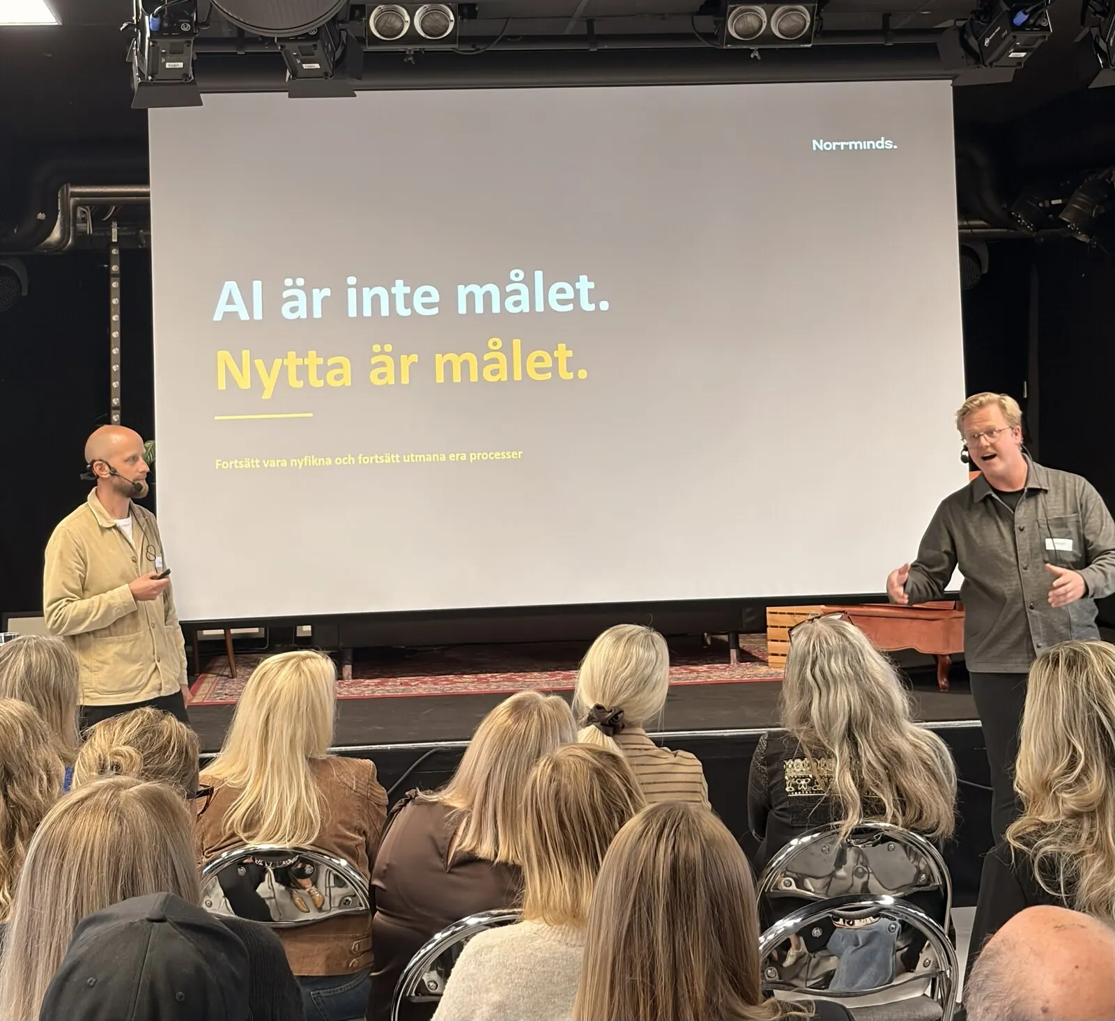 Två kollegor på scen framför en skärm med texten ”AI är inte målet. Nytta är målet.”, publiken i förgrunden.